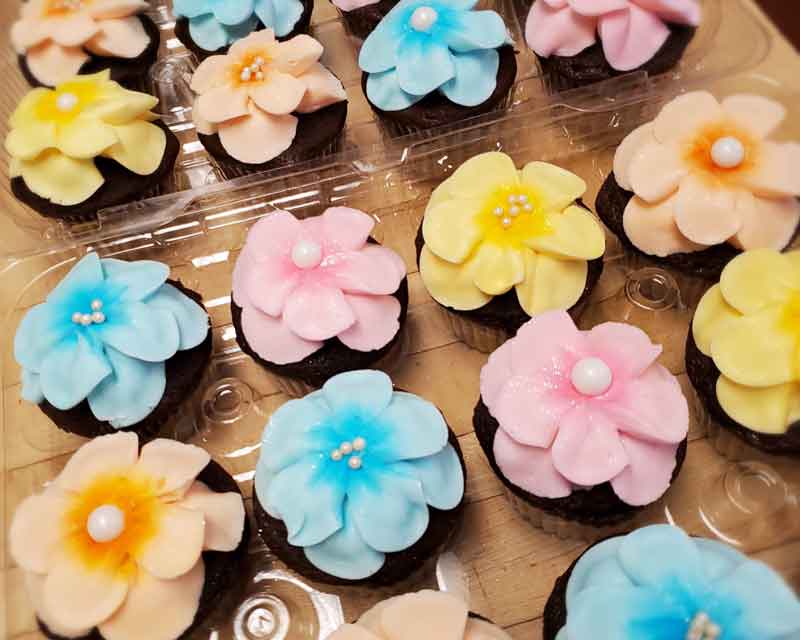A tray of chocolate cupcakes decorated with large, colorful flower-shaped frosting designs in pastel shades of blue, pink, yellow, and peach. Each flower is topped with a small edible pearl or cluster of decorative sugar beads, creating a vibrant and festive presentation.