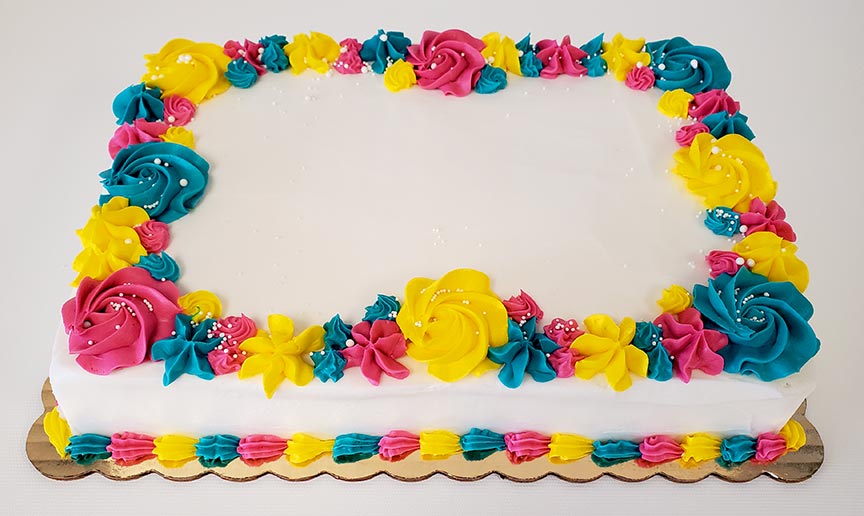 Rectangular white sheet cake with vibrant piped buttercream flowers in pink, yellow, and teal decorating the top and bottom borders. The flowers include rosettes, stars, and small blossoms with white sprinkle accents. The center of the cake is left blank for a custom message, and the cake sits on a gold scalloped cake board