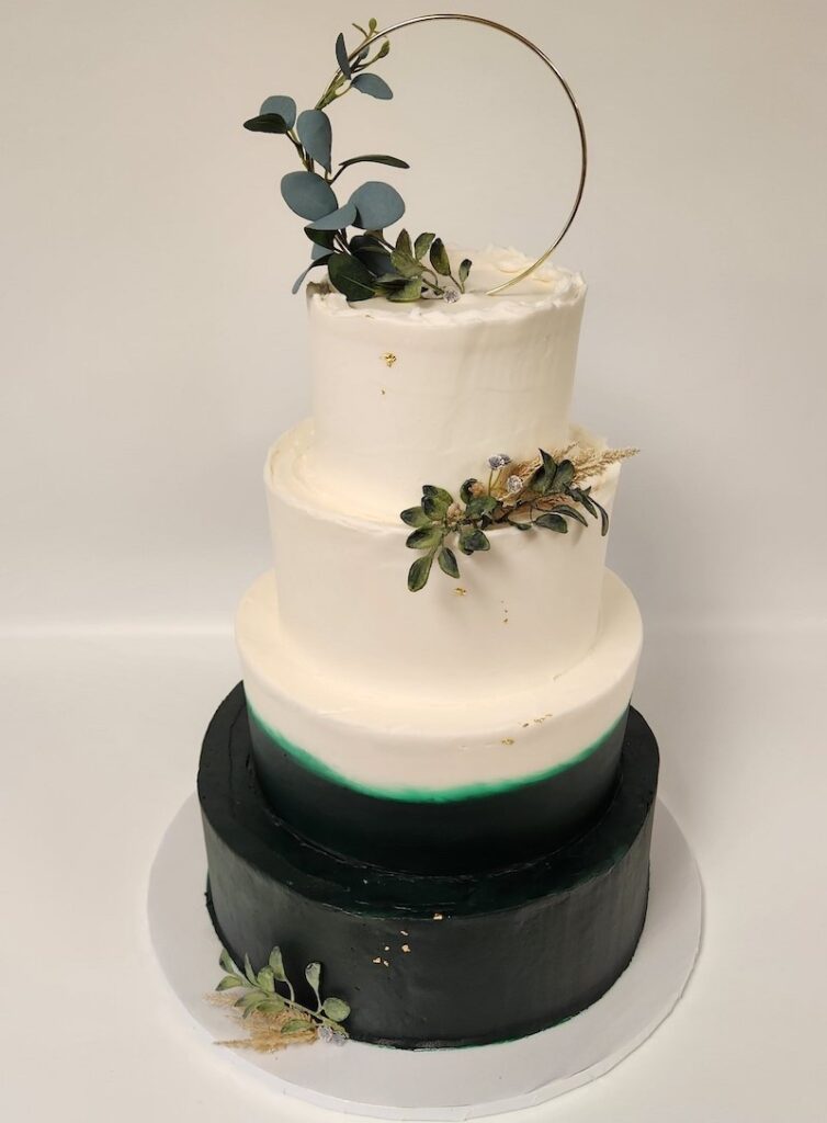 "Four-tiered wedding cake with a sleek, modern design. The top three tiers are frosted in smooth white buttercream, while the bottom tier features a glossy deep green finish with a soft gradient blending into white. The cake is decorated with clusters of green succulents, dried florals, and a gold hoop topper adorned with eucalyptus leaves. Set against a plain white background."
