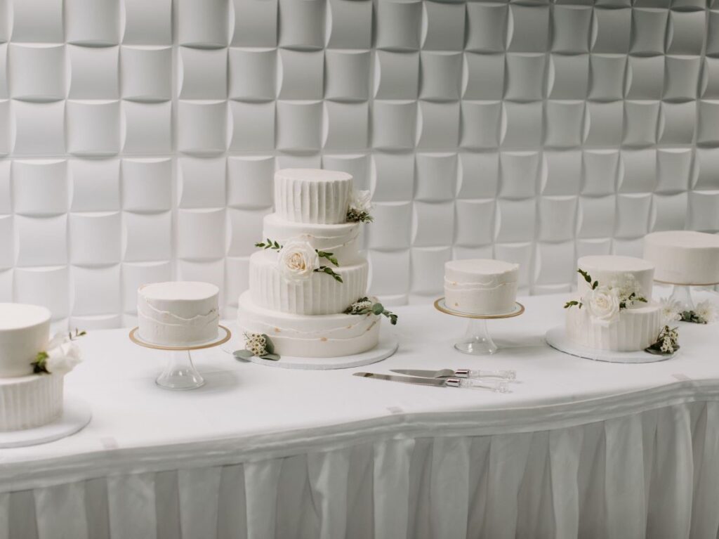 A wedding cake display featuring a central three-tiered cake surrounded by five smaller matching cakes, all decorated with white icing and delicate floral accents. The cakes are set on a white table with a pleated tablecloth, against a modern textured white wall backdrop. A cake knife and server are placed in front of the centerpiece, completing the elegant and coordinated setup.