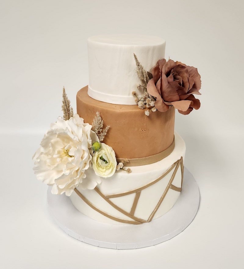 "Three-tiered wedding cake with a modern rustic design. The top tier is smooth white, the middle tier is a matte caramel brown, and the bottom tier is white with diagonal tan geometric lines. The cake is decorated with large, realistic-looking artificial flowers, including a white peony, a pale green ranunculus, and a dusty rose-colored flower, along with dried grasses and small beige accents. Set against a plain white background."