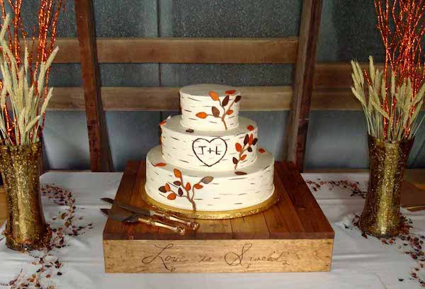 Three-tiered rustic wedding cake designed to resemble birch tree bark, with hand-painted details and a carved heart featuring the initials 'T+L.' The cake is decorated with branches and orange fondant leaves, giving it an autumn woodland feel. It sits on a wooden cake stand engraved with the phrase 'Love is Sweet.' The table is adorned with gold glitter vases filled with dried wheat and orange branches, and scattered berries, all set against a barn-style wooden backdrop