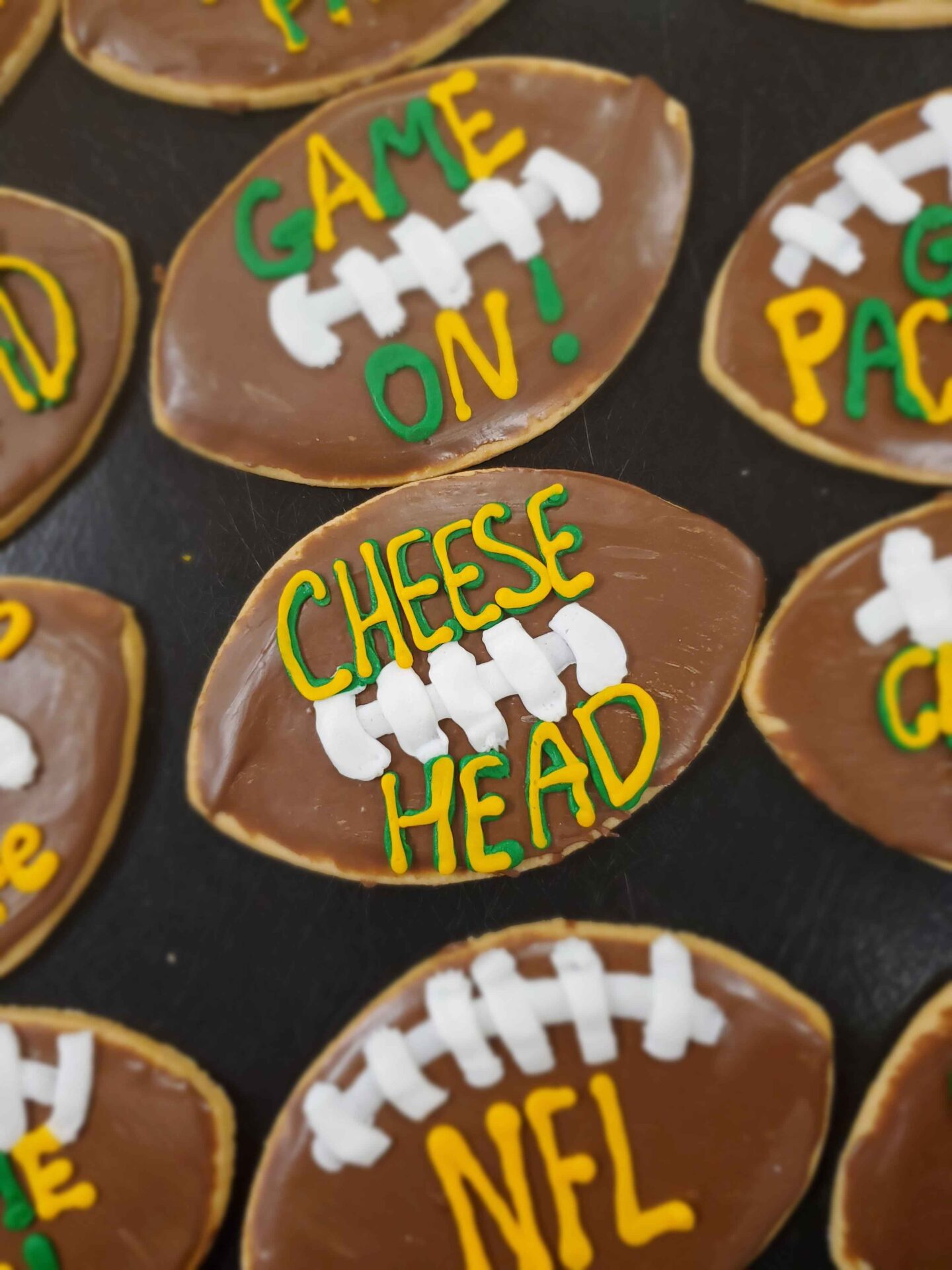 Football-shaped sugar cookies decorated with brown icing and white laces to resemble footballs. Each cookie features phrases written in green and yellow icing, including "GAME ON!", "CHEESE HEAD", and "NFL", referencing football culture and likely celebrating a game day or NFL event. The cookies are arranged on a dark surface.