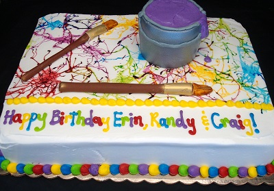 Rectangular birthday cake with a white base and a vibrant splatter-paint design on the top surface in various colors. Edible decorations include a 3D purple paint can and two realistic paintbrushes made of fondant. The front edge of the cake features a cheerful message in rainbow-colored letters reading 'Happy Birthday Erin, Randy & Craig!' The border is decorated with multicolored round candies