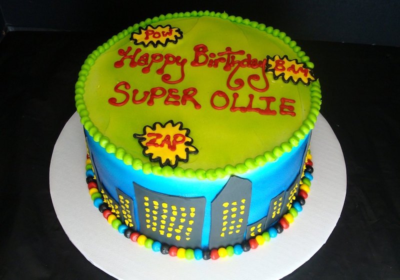 Round superhero-themed birthday cake with a lime green top and blue sides decorated to look like a city skyline with yellow-lit windows. The top features comic-style words like 'POW,' 'BAM,' and 'ZAP' in yellow explosion shapes, along with red and yellow icing text reading 'Happy Birthday Super Ollie.' The cake is bordered with a bright green bead design on top and multicolored candy beads around the base