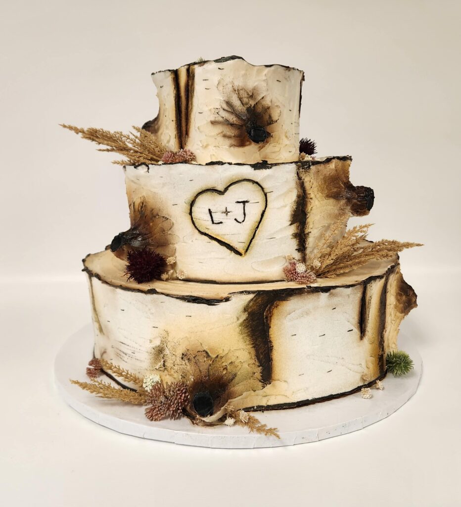Three-tiered wedding cake designed to resemble birch tree bark, complete with realistic wood grain textures and darkened edges. The middle tier features a carved heart with the initials 'L + J' inside. The cake is decorated with dried florals, including thistles and pampas grass, adding to the woodland-inspired aesthetic. Displayed against a plain white background.