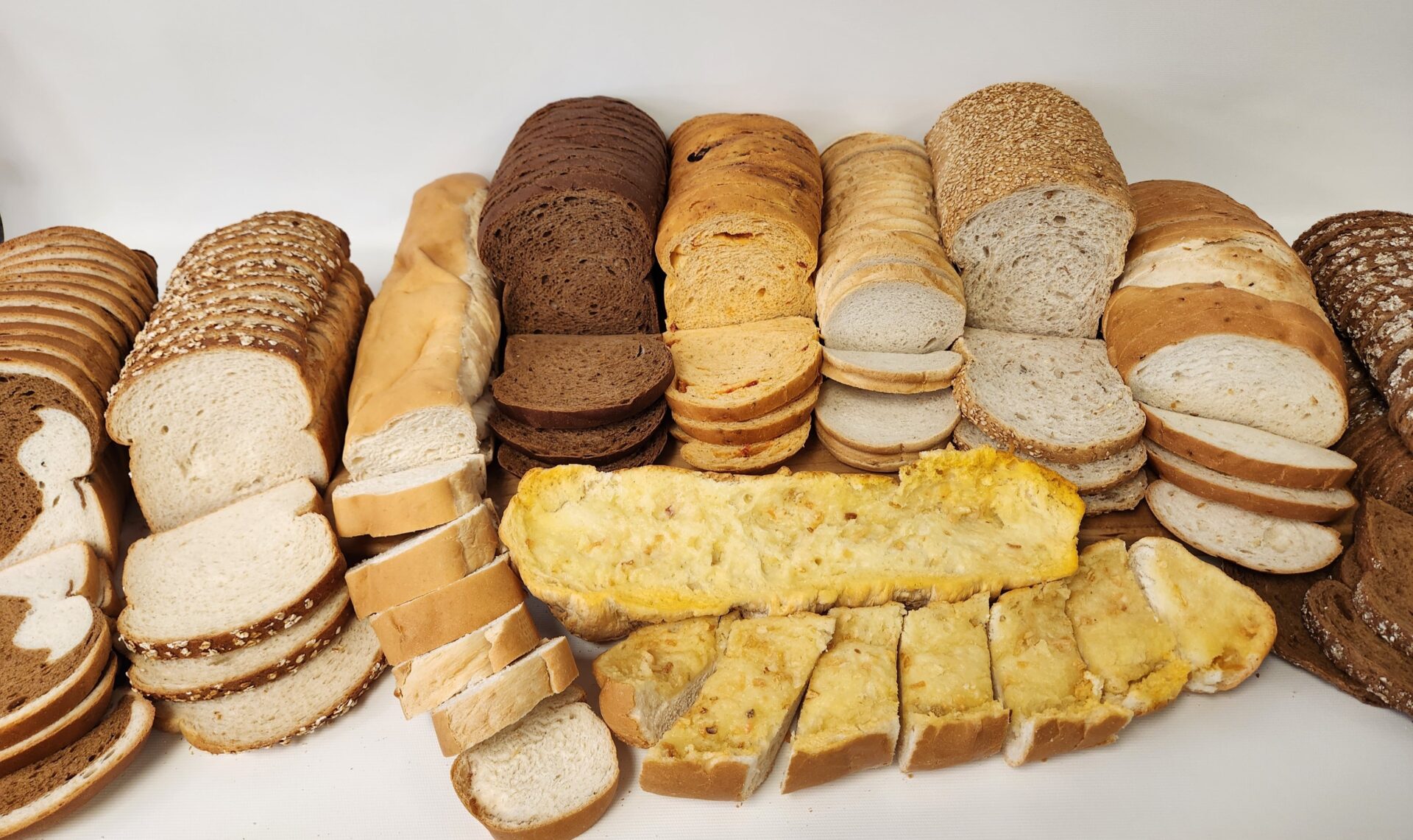 An assortment of various types of sliced bread arranged in rows against a white background. The selection includes white, wheat, rye, multigrain, and French bread, along with slices of cheese-topped and baked egg bread in the front. The display showcases the variety of textures, colors, and styles, ideal for a bakery menu or promotional image.