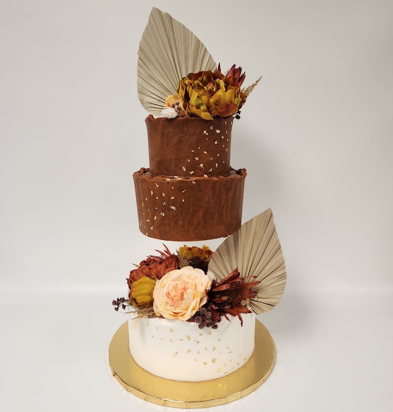 "Three-tiered cake with a rustic, modern design. The top two tiers are covered in chocolate brown frosting with scattered gold leaf accents, while the bottom tier is frosted in white with similar gold detailing. Each tier is adorned with dried floral arrangements in warm tones of burnt orange, mustard yellow, and blush, along with large, dried palm leaves. The cake is displayed on a round gold cake board against a plain white background."