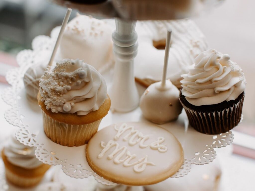 A beautifully arranged tiered dessert stand featuring elegant white and gold wedding-themed treats. Displayed are vanilla and chocolate cupcakes with swirls of white frosting, cake pops, petit fours, and a decorated sugar cookie that reads “Mr & Mrs” in cursive. The color palette is soft and romantic, ideal for a wedding or bridal celebration.