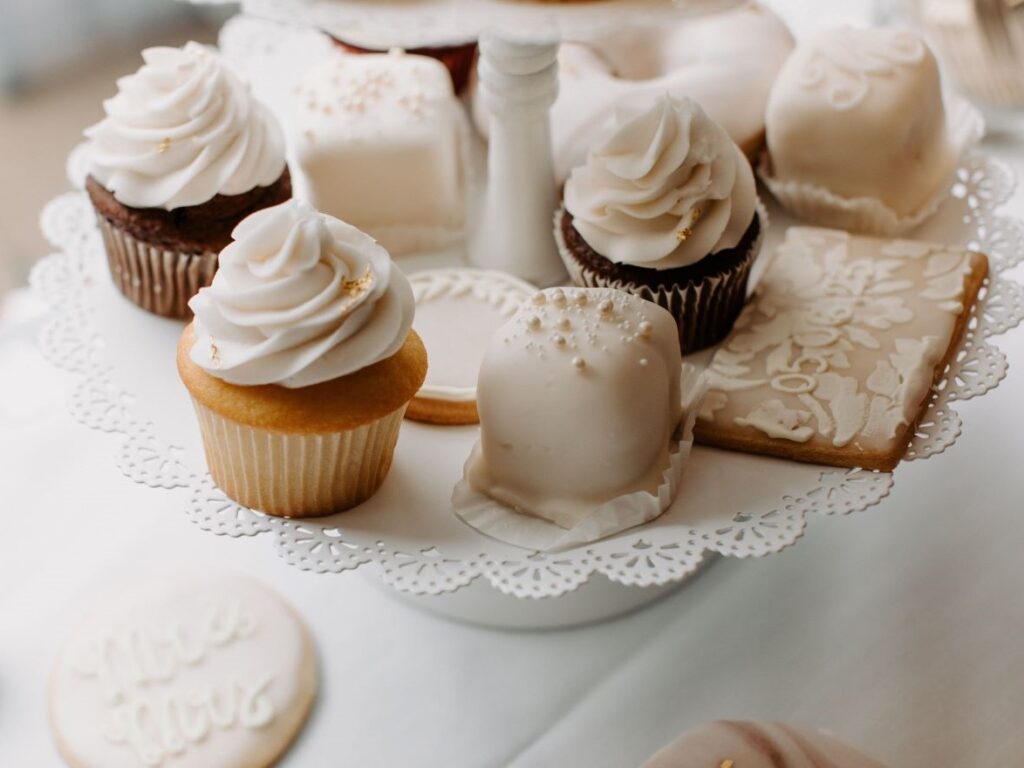 A close-up of an elegant dessert display on a white lace-trimmed cake stand, featuring vanilla and chocolate cupcakes with white frosting, petit fours, and intricately decorated sugar cookies. The treats are styled in a soft, neutral color palette with gold accents, perfect for a sophisticated wedding or bridal shower.